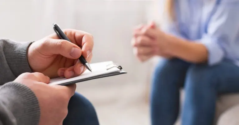 close up of a northern mindanao psychiatrist writing on a clipboard while a person sits across, hands clasped.