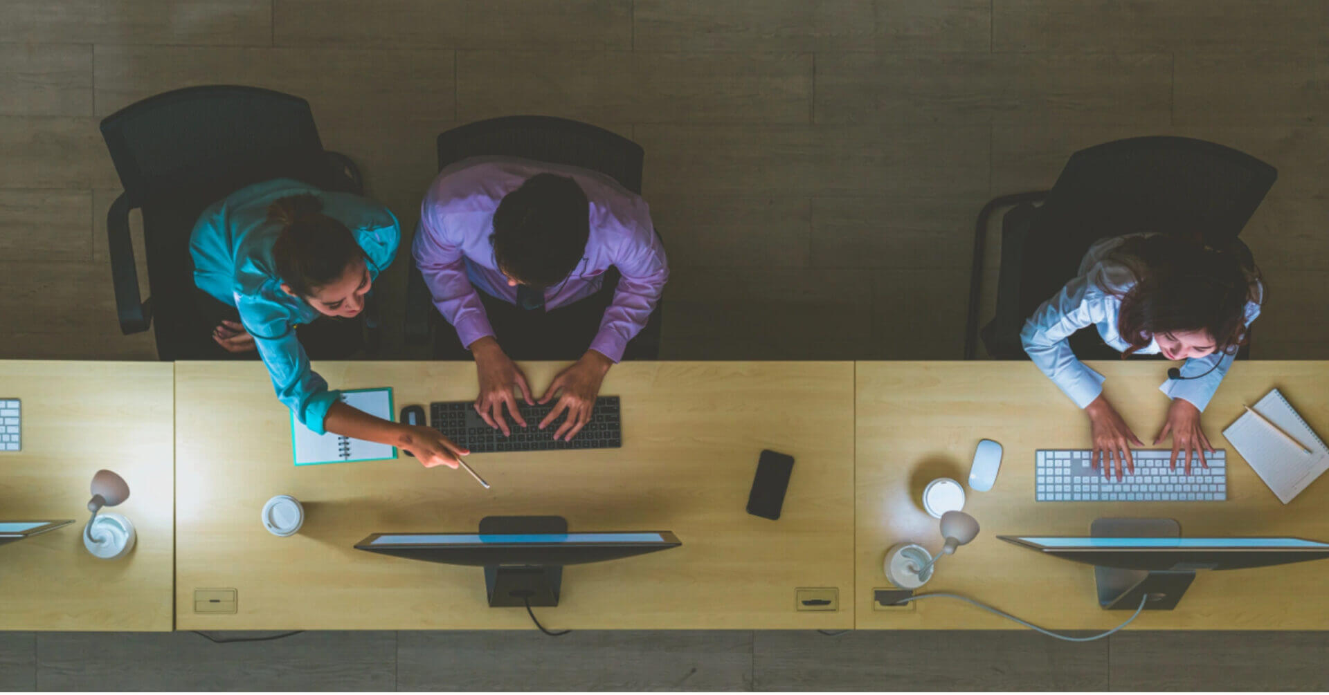three people work at computers in an iligan city office, two collaborate while one works alone, viewed from above.