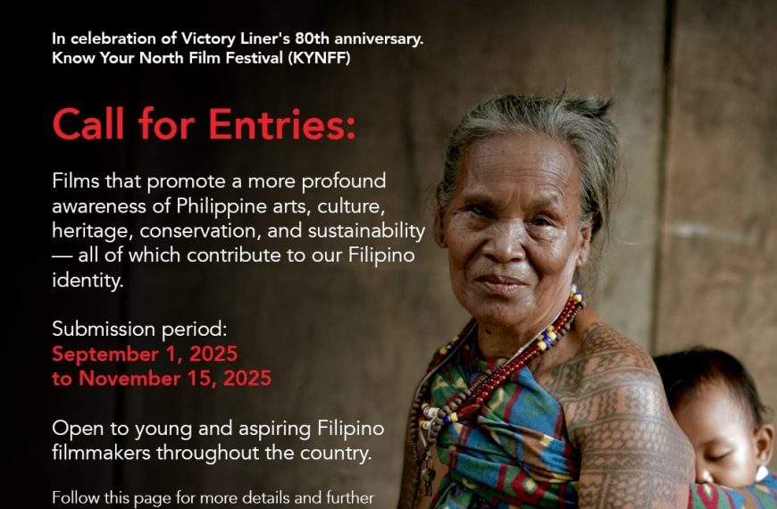 an elderly woman in traditional attire and beaded jewelry stands, arms crossed, against a blurred natural background.