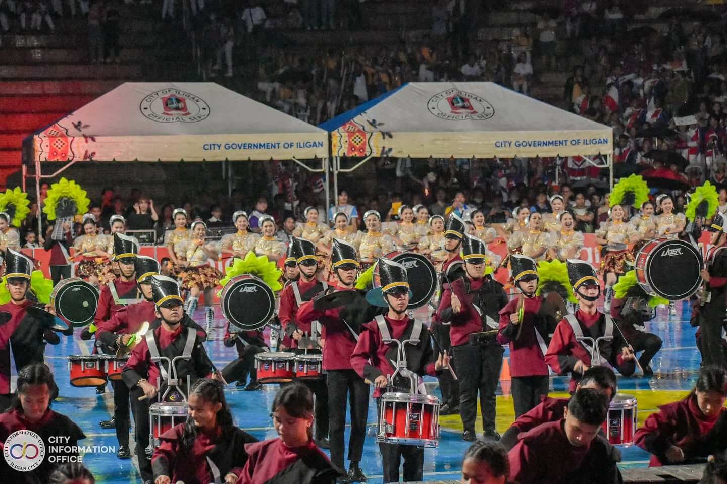 a marching band in maroon plays at the drum and lyre fest, with dancers and tents in iligan behind them.