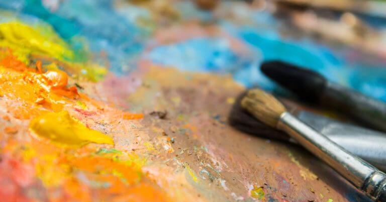 three paintbrushes rest on a messy palette at mindanao art, covered in vibrant yellow, orange, and blue paint.