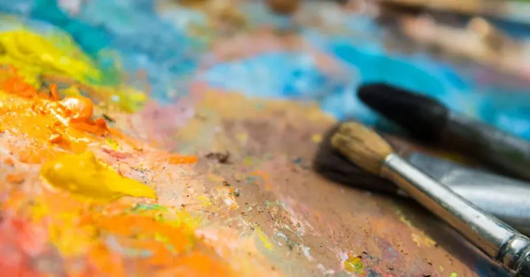 three paintbrushes rest on a messy palette at mindanao art, covered in vibrant yellow, orange, and blue paint.