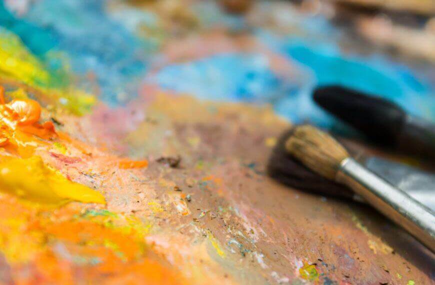 three paintbrushes rest on a messy palette at mindanao art, covered in vibrant yellow, orange, and blue paint.