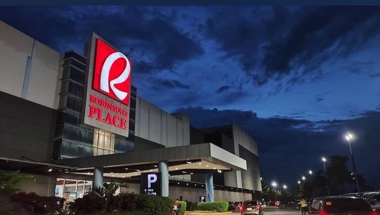 robinsons iligan mall at dusk, large red sign glowing, cars and greenery in lanao del norte’s parking area.