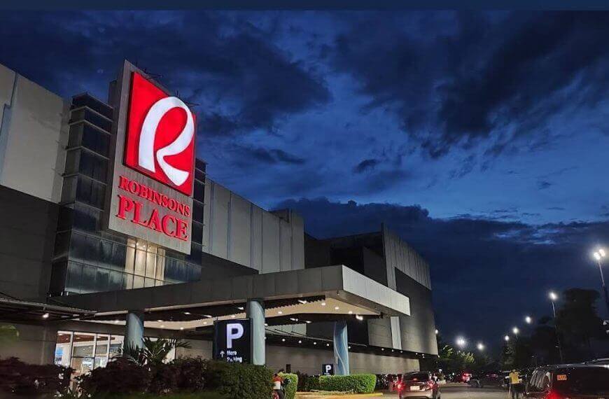 robinsons iligan mall at dusk, large red sign glowing, cars and greenery in lanao del norte’s parking area.