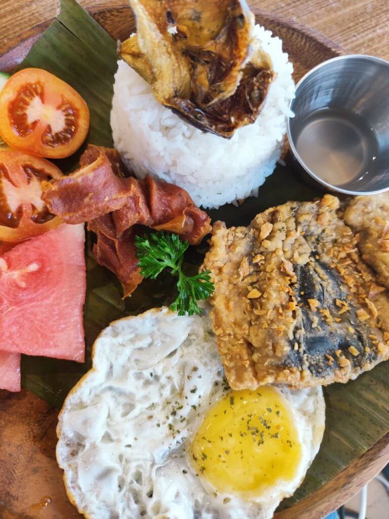 a plate with rice, fried egg, fish, bacon, tomato & watermelon at urban coffee studio’s coffee shop.