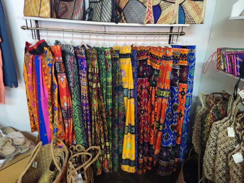 colorful patterned pants hang on a rack near iligan arts crafts, woven bags, and sandals in a store display.