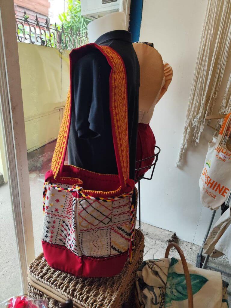 a mannequin with a black top displays an iligan arts embroidered shoulder bag with vibrant red and yellow patterns.