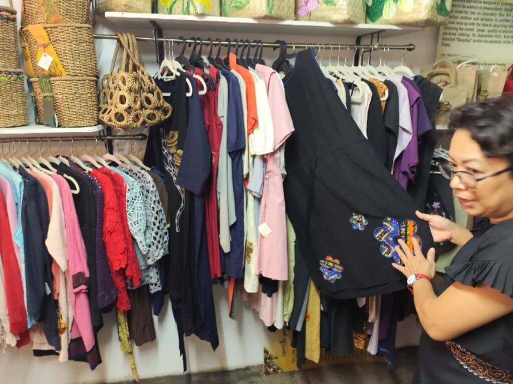 a woman browses a black embroidered dress among vibrant iligan crafts and woven bags in a lively shop.