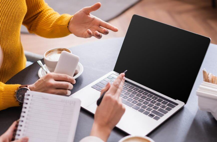 two people discuss freelancing opportunities at a table with a laptop, coffee, and phone in a cozy setting.