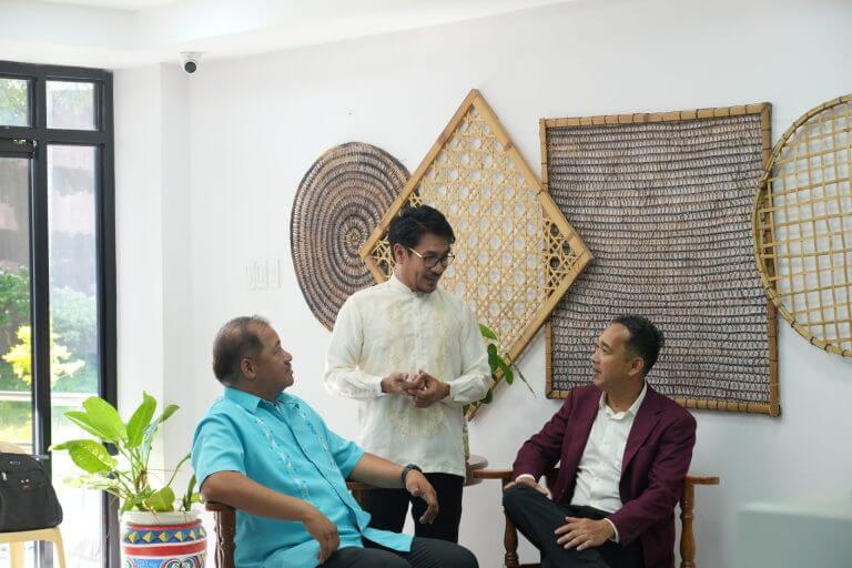 three men in dress shirts and jackets talk and smile in a bright room with woven wall decor and plants.