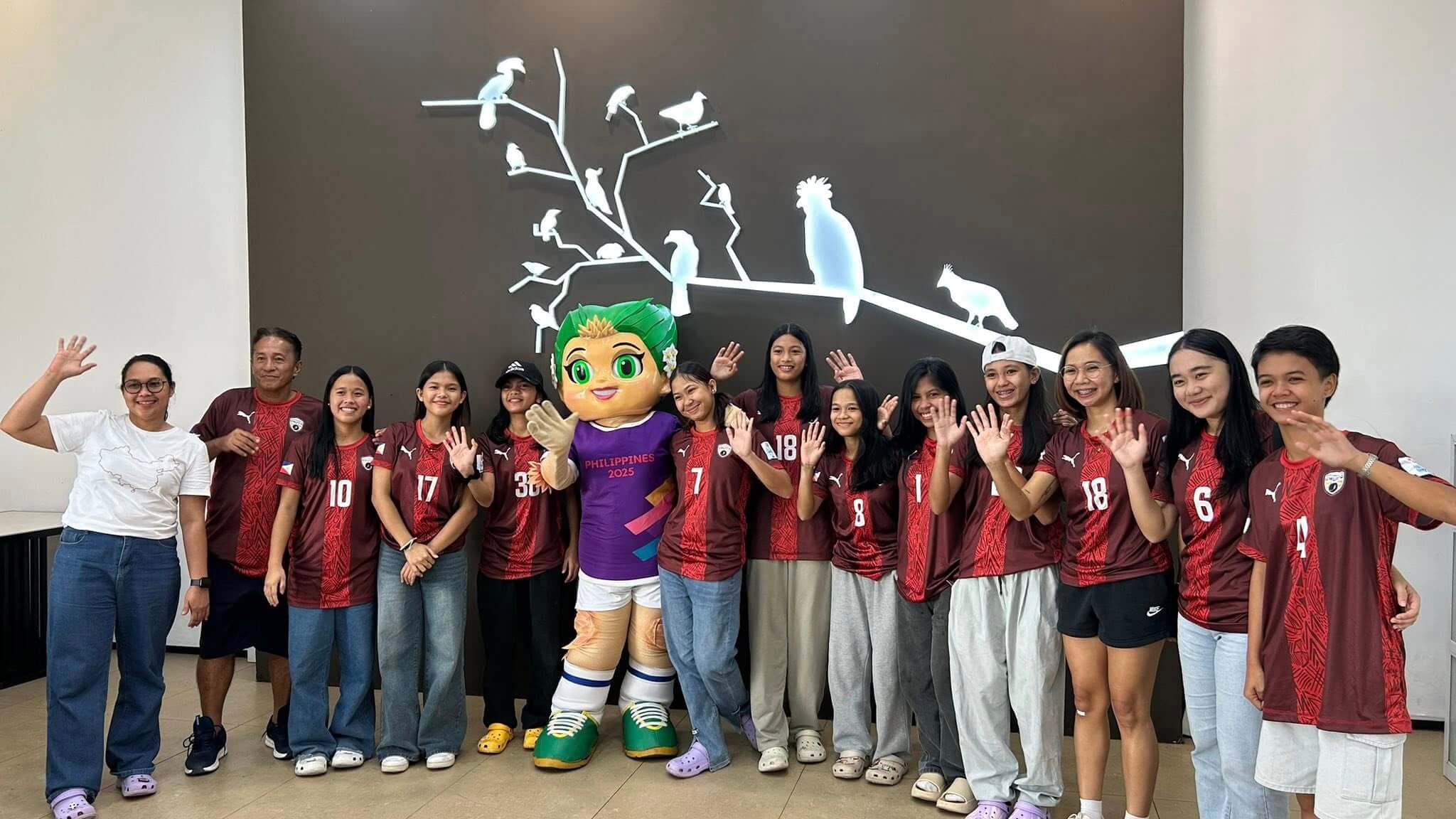 a group of people in red sports jerseys and a mascot wave, standing in front of a wall with bird silhouettes.
