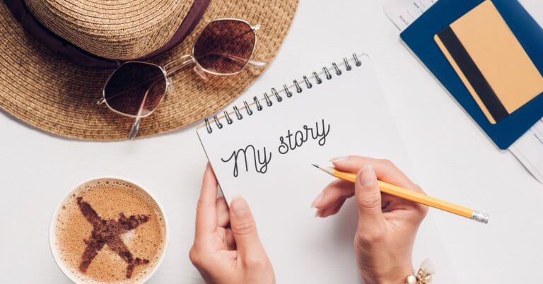 a person writes my story in a notebook beside coffee, a hat, and goiligan travel documents on a white table.