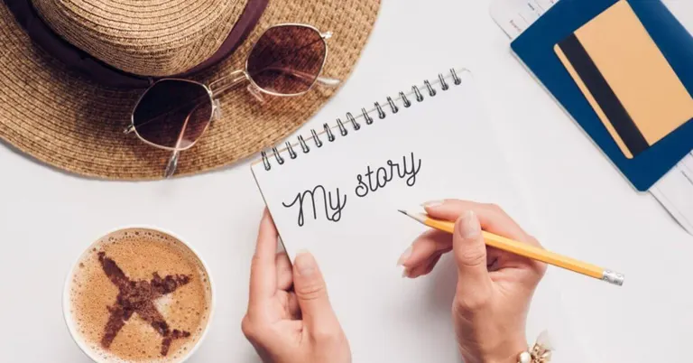 a person writes my story in a notebook beside coffee, a hat, and goiligan travel documents on a white table.
