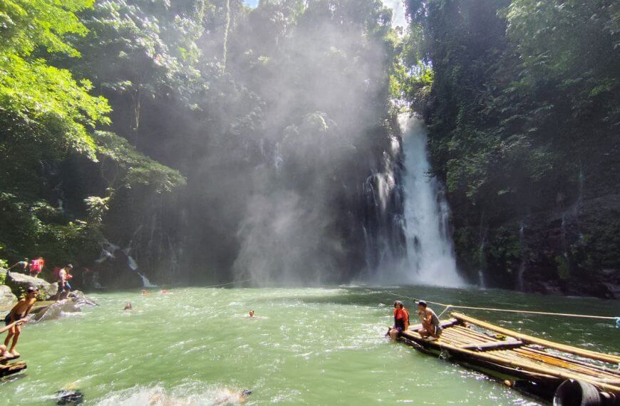 people swim and relax on bamboo rafts by iligan falls, surrounded by lush green forest in bright sunlight.