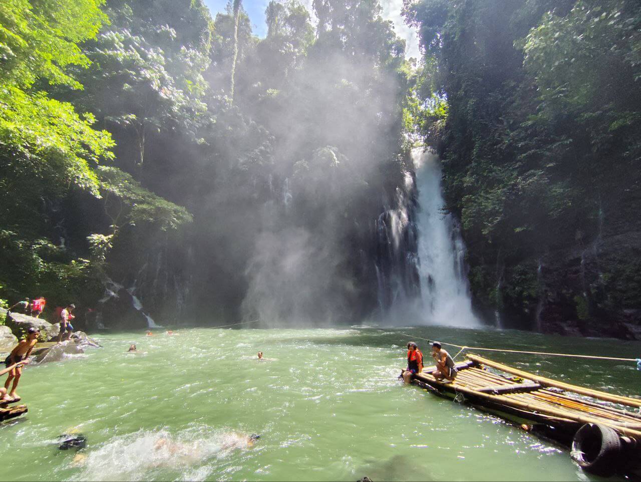 people swim and relax on bamboo rafts by iligan falls, surrounded by lush green forest in bright sunlight.