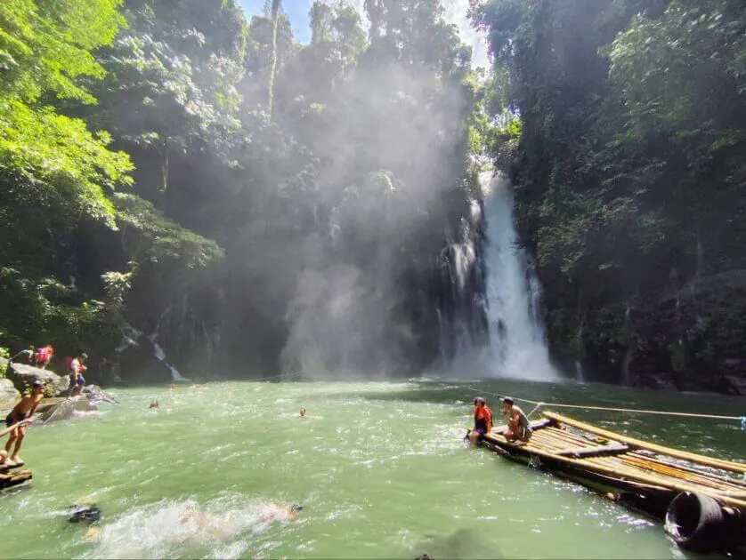 people swim and relax on bamboo rafts by iligan falls, surrounded by lush green forest in bright sunlight.
