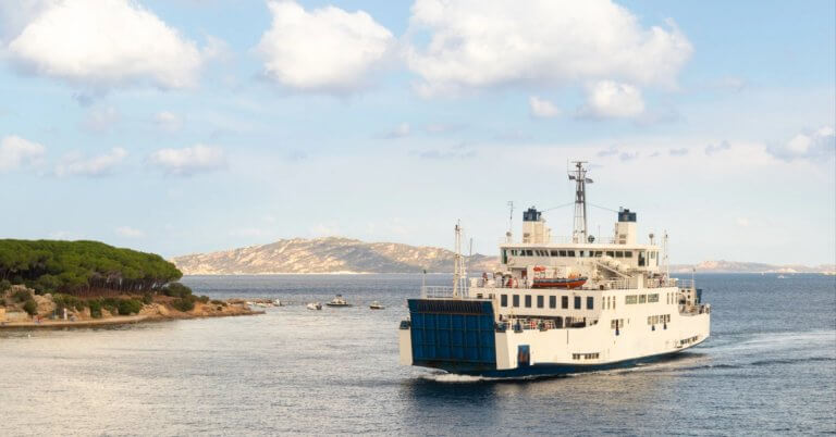 a trans asia shipping ferry sails near a tree lined coast with calm blue water and a distant hilly island.