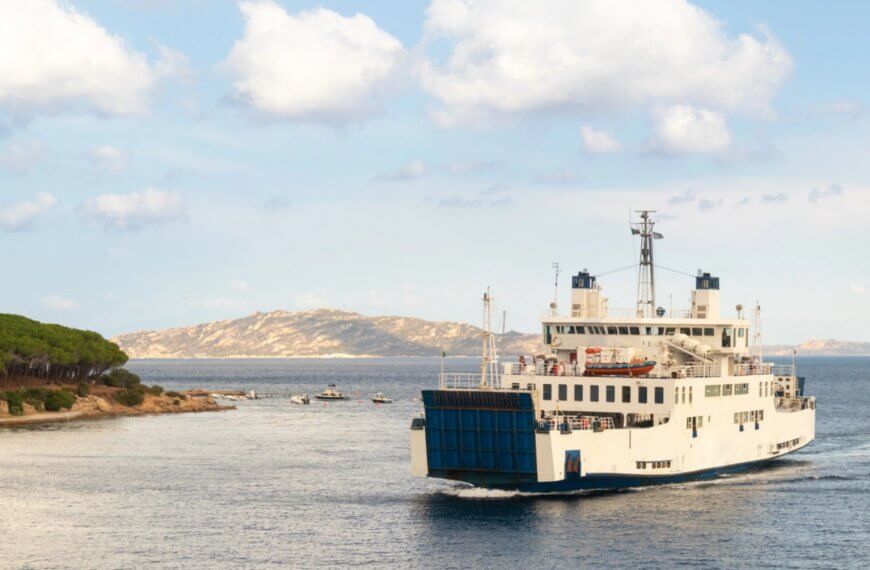 a trans asia shipping ferry sails near a tree lined coast with calm blue water and a distant hilly island.