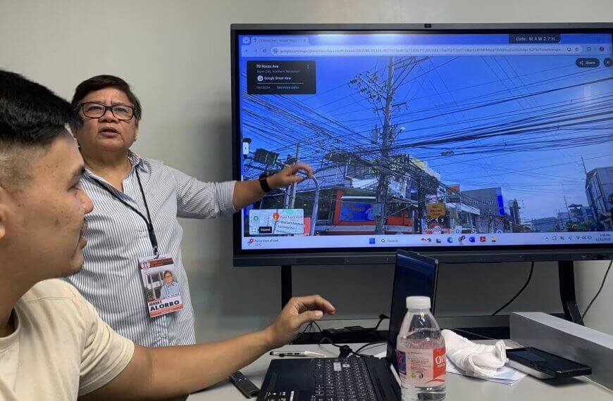 two people discuss a street view of dangling wires on a large screen in a meeting room.