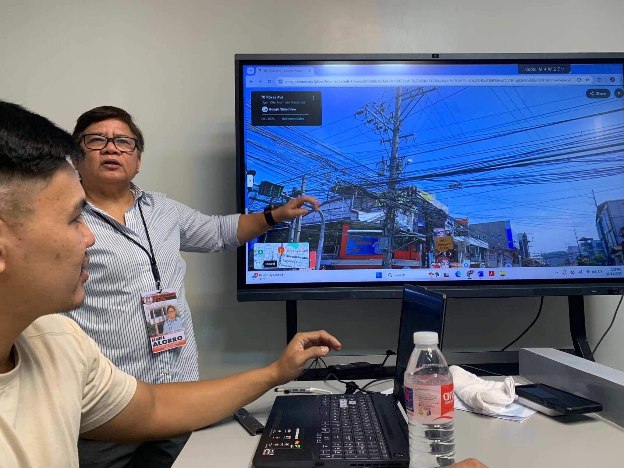 two people discuss a street view of dangling wires on a large screen in a meeting room.