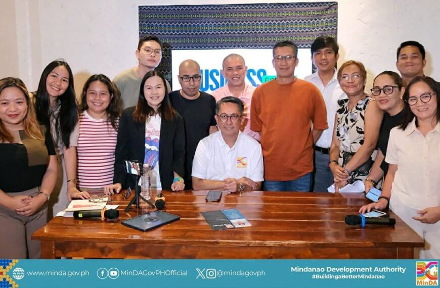 fifteen people smiling, posing together indoors behind a wooden table with microphones and papers.