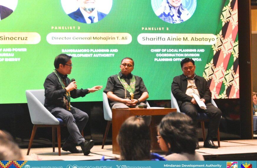 three panelists sit on stage discussing at an event, with colorful banners and a logo in the background.