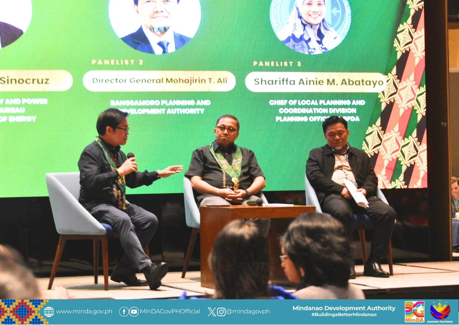 three panelists sit on stage discussing at an event, with colorful banners and a logo in the background.
