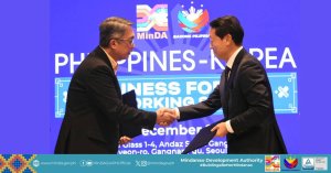 two men in suits shake hands on stage before a blue “philippines korea” business event backdrop.