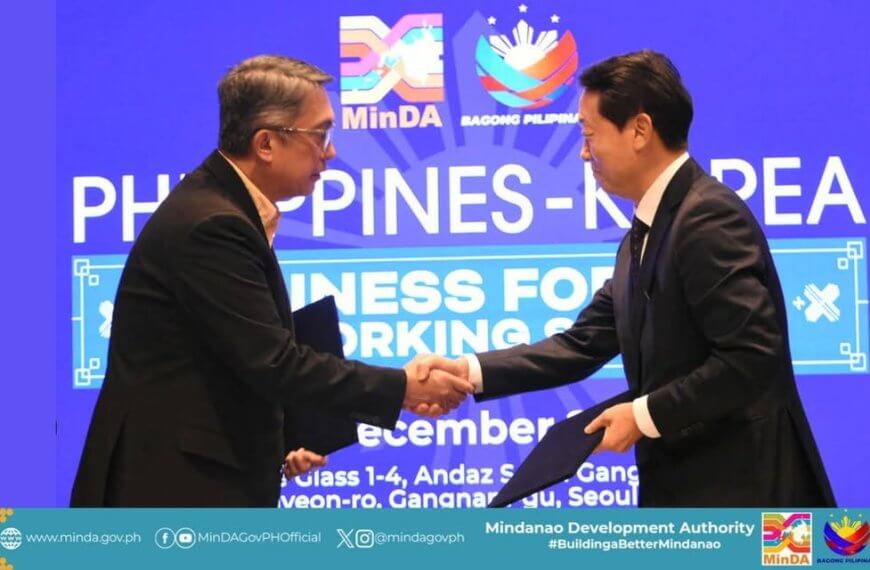 two men in suits shake hands on stage before a blue “philippines korea” business event backdrop.