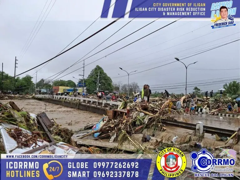 flooded road in iligan city covered with debris and damaged trees, with cloudy skies and emergency hotlines displayed.