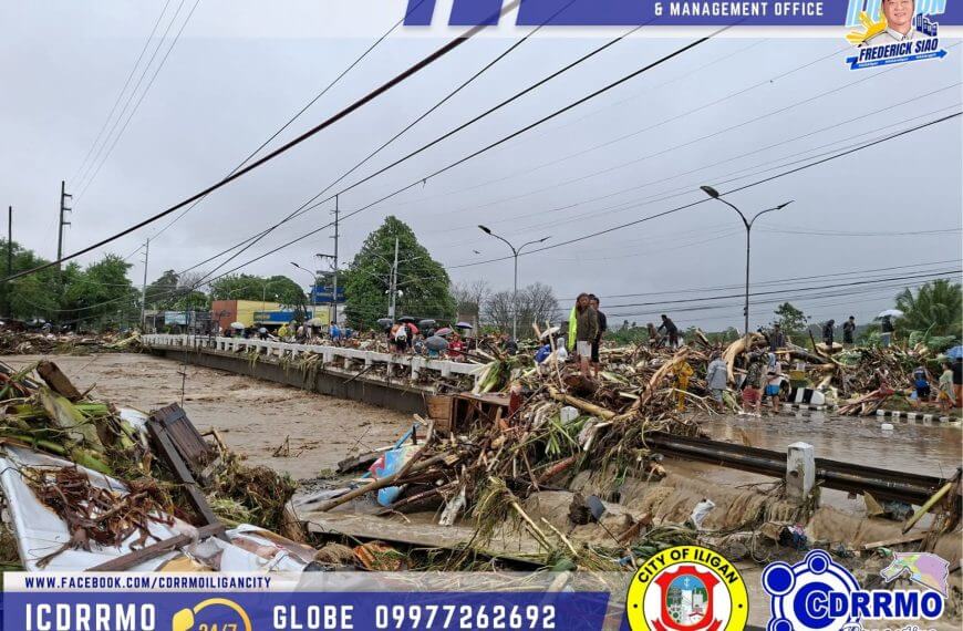 flooded road in iligan city covered with debris and damaged trees, with cloudy skies and emergency hotlines displayed.