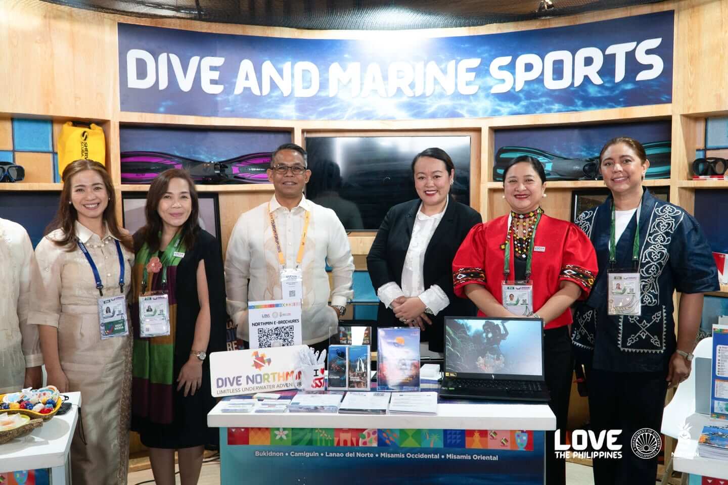six people stand smiling at a dive and marine sports booth with brochures, laptop, and colorful display.