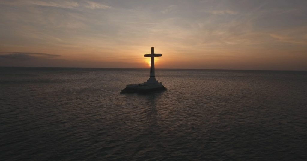 a large cross stands on a platform in the sea, silhouetted against an orange sunset&mdash;one of mindanao&rsquo;s must visit spots.