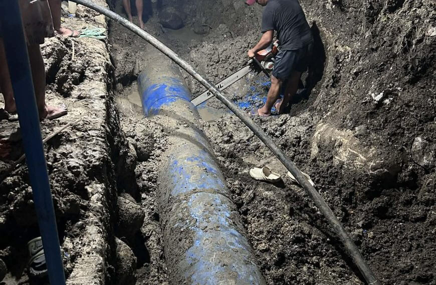 workers repair a large blue pipe in a muddy trench at night, using tools and surrounded by soil.