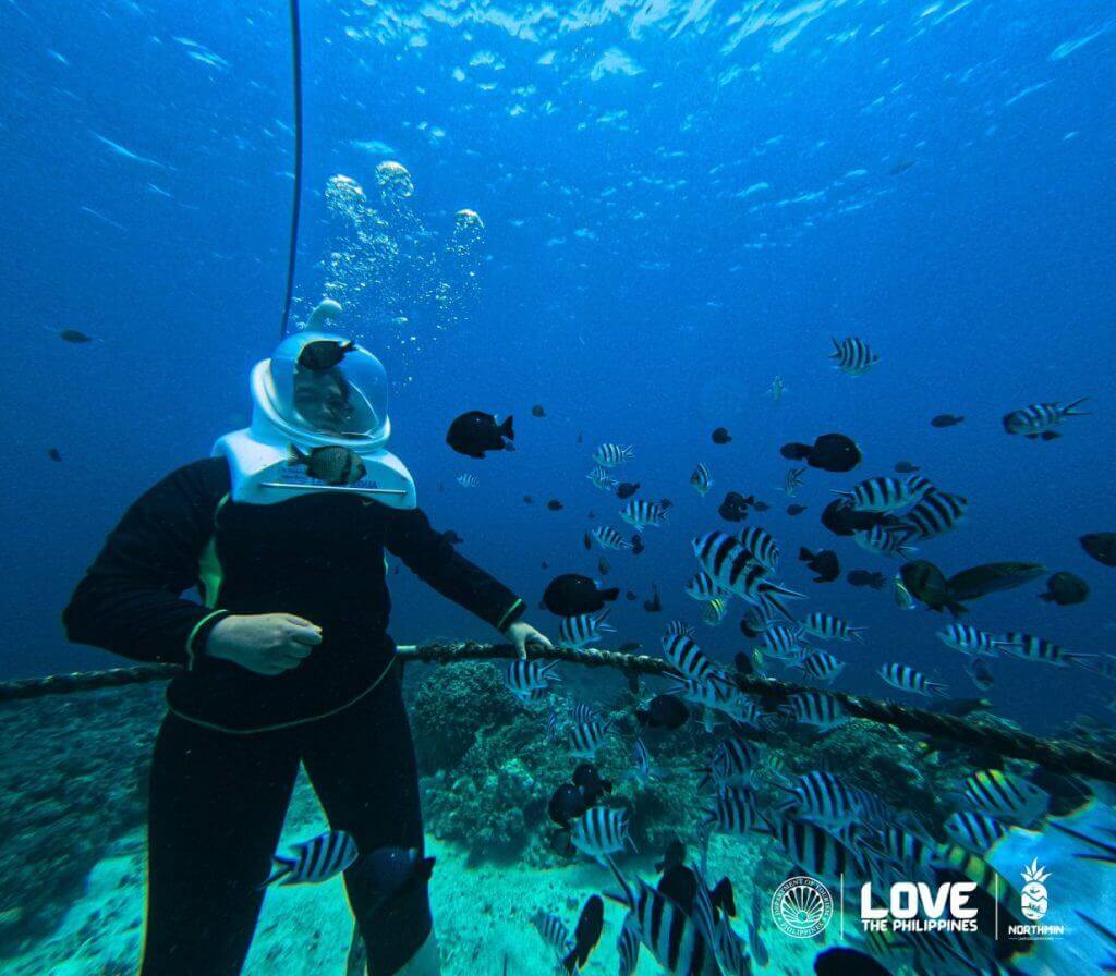 a person in a sea helmet dives among camiguin’s fish; sunken cemetery and love the philippines logos visible.